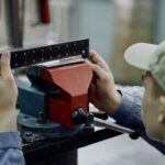 A manufacturing worker uses calipers to measure a metal component secured in a vise during quality inspection.