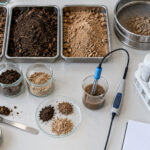 Overhead view of soil samples, a sieve, pH and conductivity probes, and preparation vessels arranged for laboratory testing